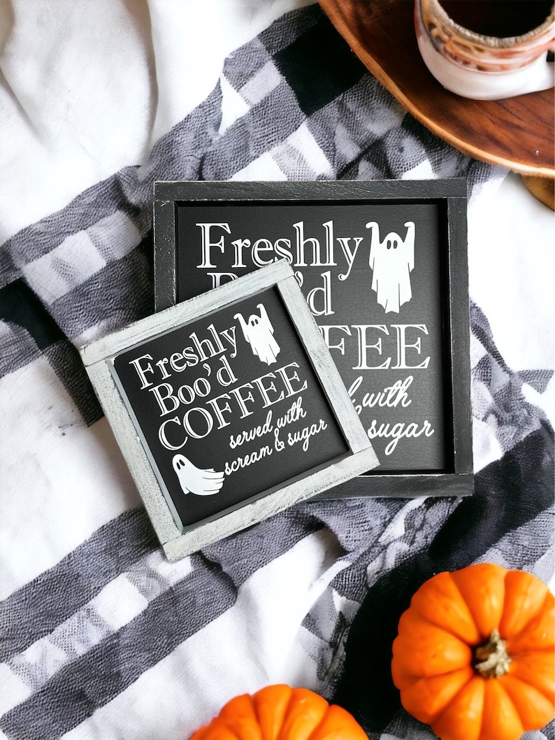 K&ouml;nnte beinhalten: Zwei schwarze gerahmte Schilder mit wei&szlig;em Text und einer Geistergrafik. Das obere Schild lautet "Freshly Boo'd Coffee" und das untere Schild lautet "serviert mit Schrei und Zucker". Die Schilder befinden sich auf einer schwarz-wei&szlig;en karierten Decke mit zwei orangefarbenen K&uuml;rbissen.