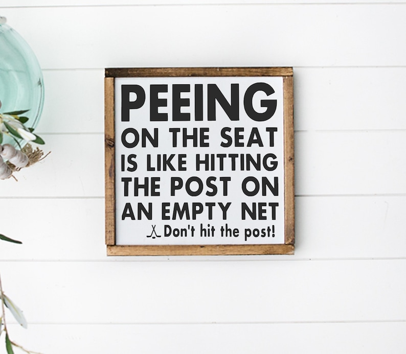 May include: A white wooden sign with a brown frame, featuring the text "PEEING ON THE SEAT IS LIKE HITTING THE POST ON AN EMPTY NET Don't hit the post!"