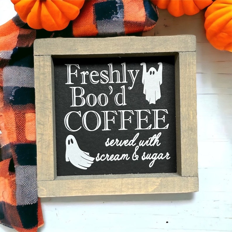 K&ouml;nnte beinhalten: Ein Holzschild mit schwarzem Hintergrund und wei&szlig;em Text, der "Freshly Boo'd Coffee served with scream & sugar" lautet. Auf dem Schild sind zwei wei&szlig;e Geister-Silhouetten zu sehen. Das Schild ist in einen hellbraunen Holzrahmen gefasst.