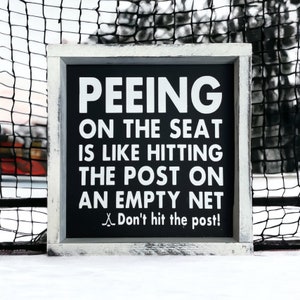 May include: A black framed sign with white text that reads "Peeing on the seat is like hitting the post on an empty net. Don't hit the post!" The sign is sitting on a white surface.