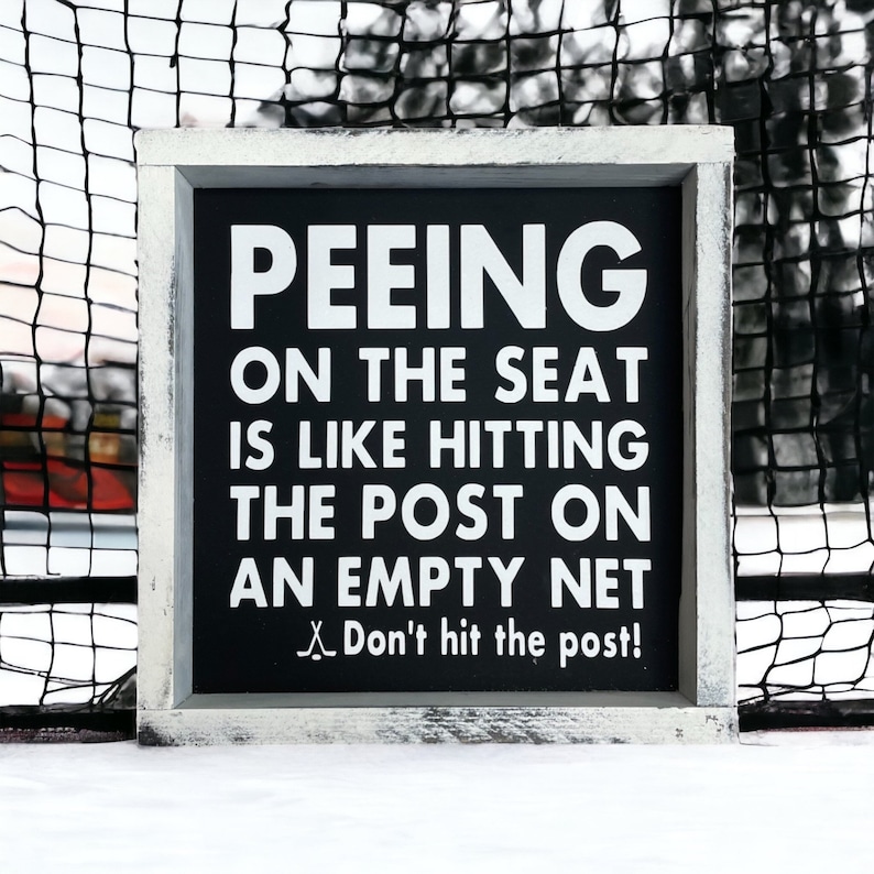May include: A black framed sign with white text that reads "Peeing on the seat is like hitting the post on an empty net. Don't hit the post!" The sign is sitting on a white surface.