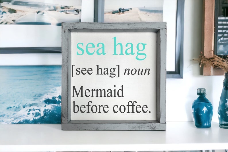 K&ouml;nnte beinhalten: Ein gerahmtes Schild mit dem Text "sea hag [see hag] noun Mermaid before coffee." Das Schild ist in hellgrau gestrichen und hat einen wei&szlig;en Hintergrund.