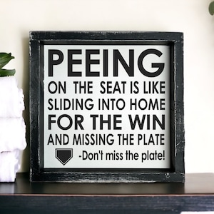 May include: A black framed sign with white text that reads "Peeing on the seat is like sliding into home for the win and missing the plate - Don't miss the plate!" The sign has a black baseball home plate graphic in the bottom left corner.