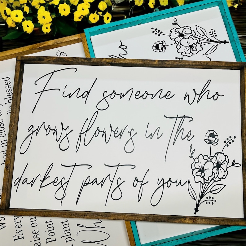 May include: White wooden sign with black calligraphy that reads "Find someone who grows flowers in the darkest parts of you". The sign is decorated with a black floral design.
