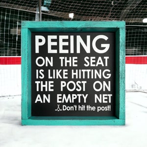 May include: A teal framed sign with a black background and white text that reads "PEEING ON THE SEAT IS LIKE HITTING THE POST ON AN EMPTY NET. Don't hit the post!"