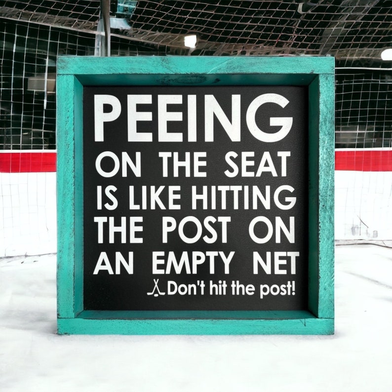 May include: A teal framed sign with a black background and white text that reads "PEEING ON THE SEAT IS LIKE HITTING THE POST ON AN EMPTY NET. Don't hit the post!"