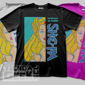 Puede incluir: Tres camisetas en blanco, negro y morado, cada una con un gráfico retro de una mujer rubia con ojos azules. Los textos "PRINCESS OF POWER" y "SHE-RA" están impresos en las camisetas.