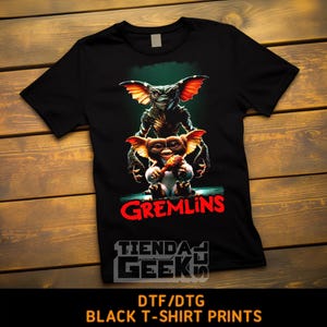 Może przedstawiać: Czarny t-shirt z kolorową grafiką przedstawiającą dwie postacie Gremlinów. Słowo "Gremlins" jest wydrukowane na czerwono, poniżej znajduje się tekst "TIENDA GEEKS". Na dole koszulki widnieje napis "DTF/DTG BLACK T-SHIRT PRINTS".