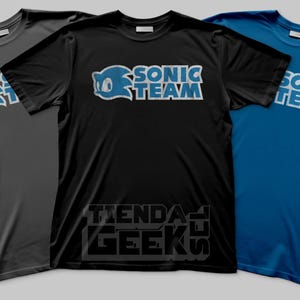 Puede incluir: Tres camisetas en gris, negro y azul. Cada camiseta presenta el texto "SONIC TEAM" en blanco y azul, junto con un personaje de dibujos animados azul. La camiseta negra tiene el texto "TIENDA GEEKS" en la parte inferior.