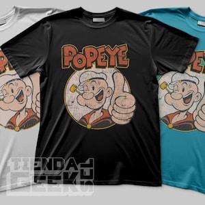 Puede incluir: Tres camisetas en blanco, negro y verde azulado, cada una con un gráfico de estilo vintage de Popeye el Marino. El diseño incluye la imagen de Popeye con el pulgar hacia arriba y la palabra "Popeye" en letras naranjas.
