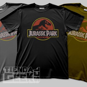 Puede incluir: Tres camisetas en gris, negro y verde oliva, cada una con el logo de "Jurassic Park". El logo incluye una silueta de dinosaurio dentro de un círculo rojo y el texto "Jurassic Park" en un marco rectangular.