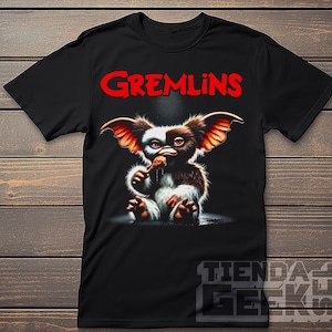 May include: Black t-shirt featuring a graphic of a Gremlin eating food, with the word "Gremlins" in red above the image. The Gremlin has brown, white, and black fur, with large ears and is holding food in its hands.