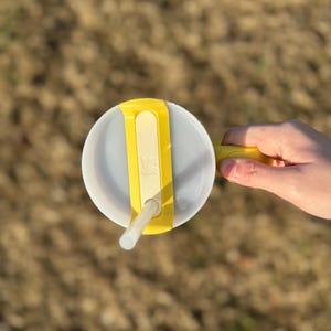 Puede incluir: Un vaso de pl&aacute;stico transparente con tapa y asa amarillas, y una pajita blanca. La tapa tiene una correa amarilla para sujetar la pajita. El vaso est&aacute; sujeto por una mano.