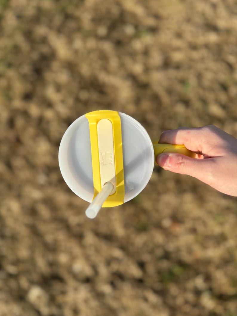 Puede incluir: Un vaso de pl&aacute;stico transparente con tapa y asa amarillas, y una pajita blanca. La tapa tiene una correa amarilla para sujetar la pajita. El vaso est&aacute; sujeto por una mano.