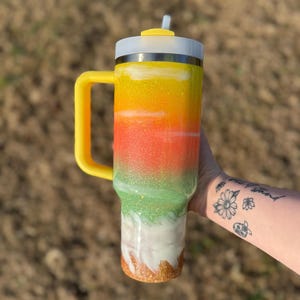 Puede incluir: Un vaso colorido con asa y tapa amarillas, y una pajita. El vaso presenta un dise&ntilde;o degradado con purpurina amarilla, naranja, verde, blanca y dorada. El vaso est&aacute; dise&ntilde;ado para mantener las bebidas fr&iacute;as o calientes.