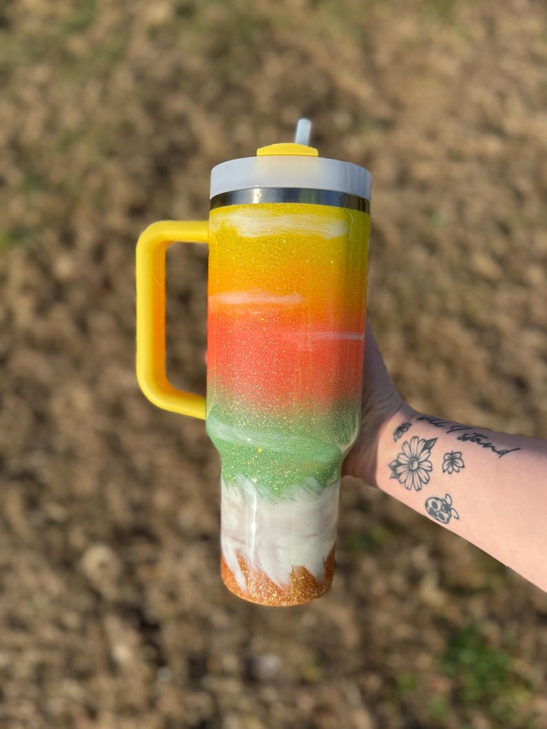 Puede incluir: Un vaso colorido con asa y tapa amarillas, y una pajita. El vaso presenta un dise&ntilde;o degradado con purpurina amarilla, naranja, verde, blanca y dorada. El vaso est&aacute; dise&ntilde;ado para mantener las bebidas fr&iacute;as o calientes.