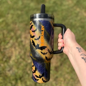 Puede incluir: Un vaso Stanley negro y dorado con asa, con un dise&ntilde;o de silueta de murci&eacute;lago. El vaso tiene una tapa negra y una pajita. La palabra "STANLEY" est&aacute; impresa en naranja en el lateral.