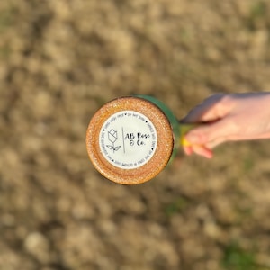 Puede incluir: Un vaso verde y dorado con purpurina y una base redonda y texturizada. La base presenta una pegatina blanca con el texto "AB Rose & Co." y un dise&ntilde;o floral. El vaso est&aacute; sostenido por una persona.