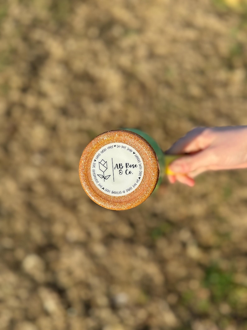 Puede incluir: Un vaso verde y dorado con purpurina y una base redonda y texturizada. La base presenta una pegatina blanca con el texto "AB Rose & Co." y un dise&ntilde;o floral. El vaso est&aacute; sostenido por una persona.