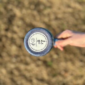 Puede incluir: Un vaso azul brillante con el texto "AB Rose & Co." e instrucciones de cuidado. Se ve la parte inferior del vaso, sostenida por una mano, con un fondo borroso de hierba.