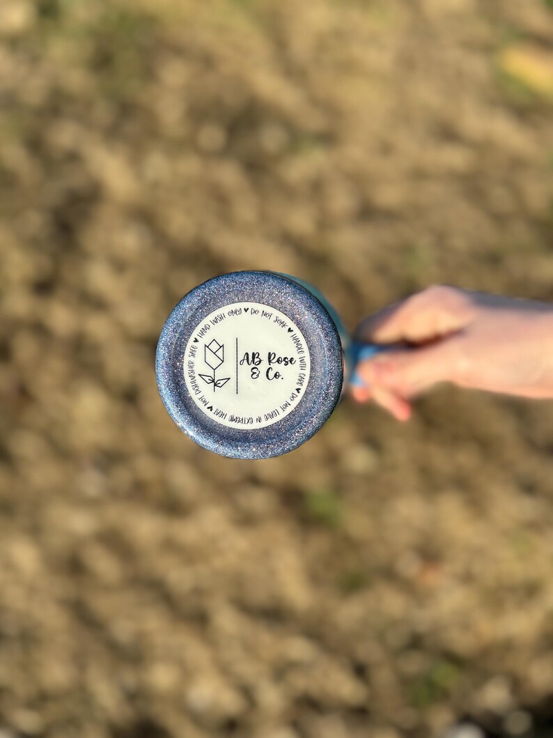 Puede incluir: Un vaso azul brillante con el texto "AB Rose & Co." e instrucciones de cuidado. Se ve la parte inferior del vaso, sostenida por una mano, con un fondo borroso de hierba.