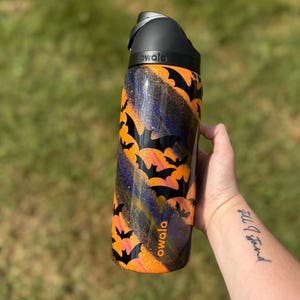 Puede incluir: Una botella de agua negra y naranja con un dise&ntilde;o de murci&eacute;lagos. La botella tiene una tapa negra y la marca "owala" est&aacute; impresa en el lateral. El fondo es verde borroso.