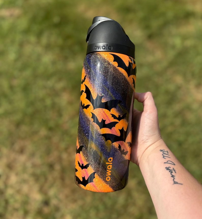 Puede incluir: Una botella de agua negra y naranja con un dise&ntilde;o de murci&eacute;lagos. La botella tiene una tapa negra y la marca "owala" est&aacute; impresa en el lateral. El fondo es verde borroso.