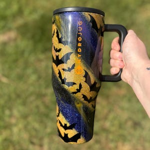 Puede incluir: Un gran termo negro con asa y tapa negras. La taza est&aacute; decorada con murci&eacute;lagos dorados con purpurina y un patr&oacute;n arremolinado de purpurina dorada, negra y azul. La palabra "HYDROJUG" est&aacute; impresa verticalmente en el lateral.