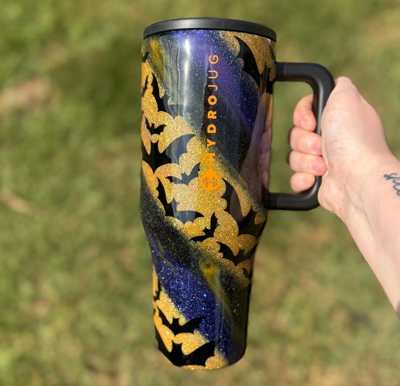 Puede incluir: Un gran termo negro con asa y tapa negras. La taza est&aacute; decorada con murci&eacute;lagos dorados con purpurina y un patr&oacute;n arremolinado de purpurina dorada, negra y azul. La palabra "HYDROJUG" est&aacute; impresa verticalmente en el lateral.