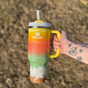 Puede incluir: Un vaso Stanley colorido con asa y tapa amarillas, y una pajita blanca. El vaso presenta un dise&ntilde;o degradado con purpurina amarilla, naranja, verde, blanca y dorada. El logotipo de Stanley es visible.