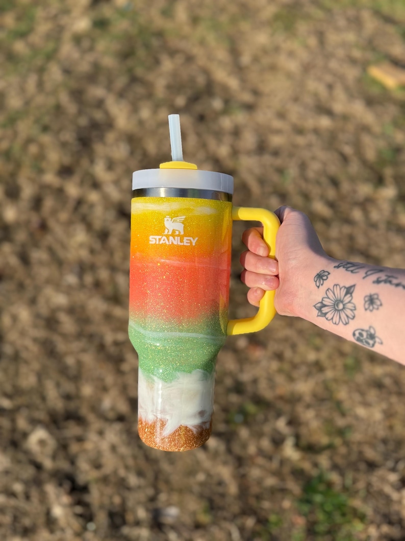 Puede incluir: Un vaso Stanley colorido con asa y tapa amarillas, y una pajita blanca. El vaso presenta un dise&ntilde;o degradado con purpurina amarilla, naranja, verde, blanca y dorada. El logotipo de Stanley es visible.