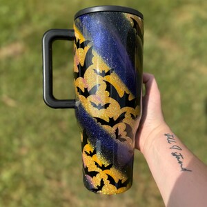 Puede incluir: Un vaso grande con asa y tapa negras. El vaso presenta un dise&ntilde;o de murci&eacute;lagos dorados con purpurina sobre un fondo de purpurina negra, morada y dorada. Dise&ntilde;ado para bebidas fr&iacute;as.