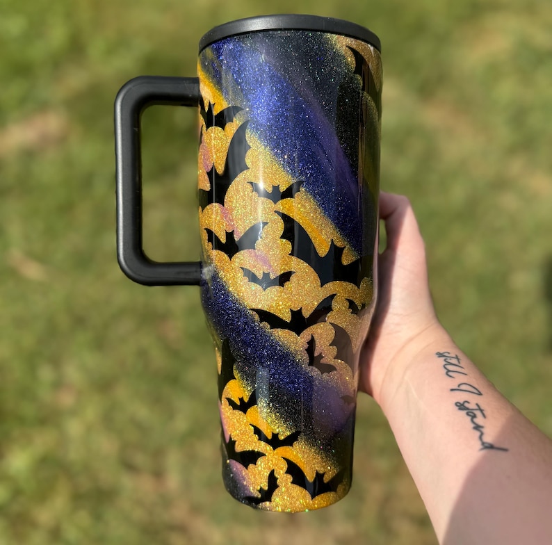 Puede incluir: Un vaso grande con asa y tapa negras. El vaso presenta un dise&ntilde;o de murci&eacute;lagos dorados con purpurina sobre un fondo de purpurina negra, morada y dorada. Dise&ntilde;ado para bebidas fr&iacute;as.