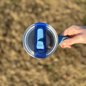 Puede incluir: Una tapa de botella de agua de pl&aacute;stico transparente con un cierre abatible azul y un asa azul. La tapa es redonda y tiene un centro transparente, lo que permite ver el contenido de la botella. La botella la sostiene una persona.