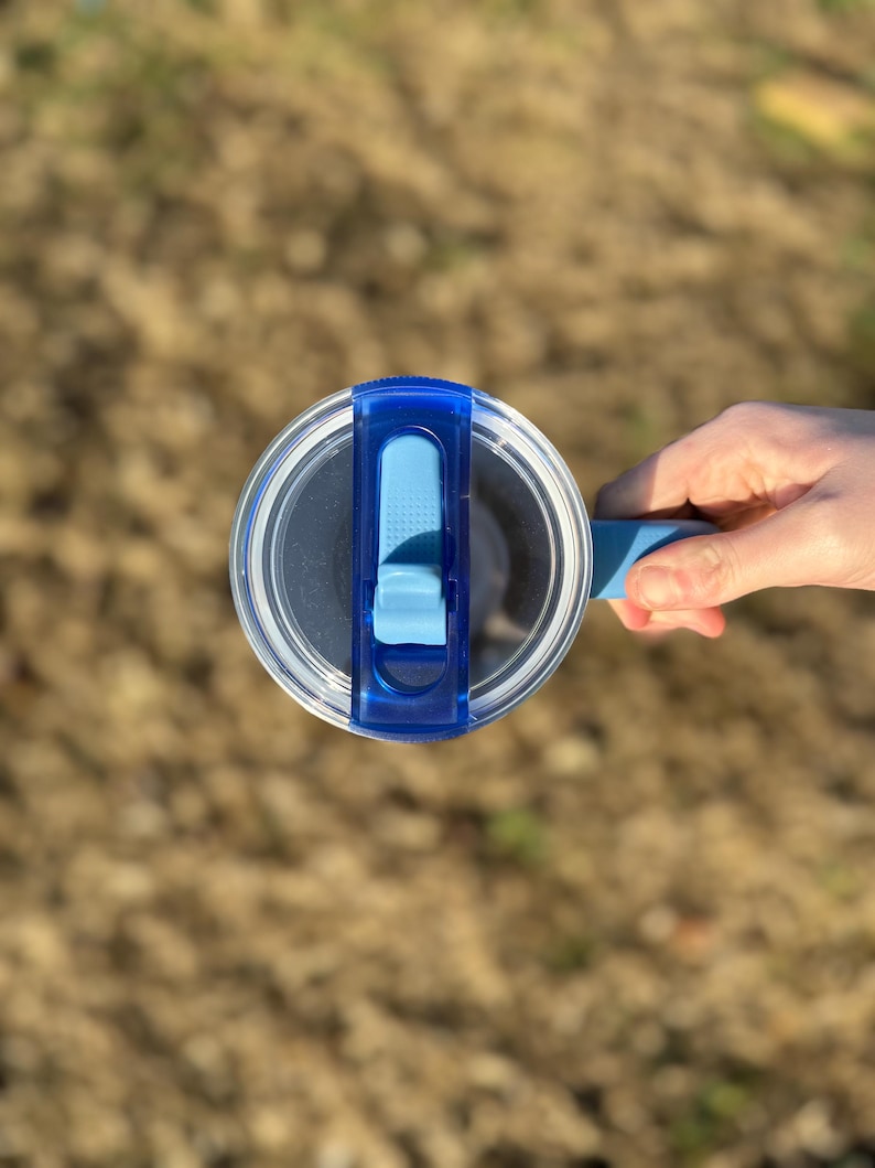 Puede incluir: Una tapa de botella de agua de pl&aacute;stico transparente con un cierre abatible azul y un asa azul. La tapa es redonda y tiene un centro transparente, lo que permite ver el contenido de la botella. La botella la sostiene una persona.