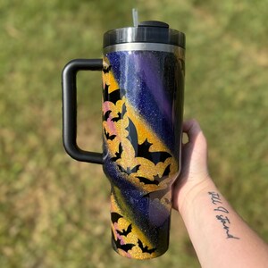 Puede incluir: Un vaso grande y aislado con asa y tapa negras. El vaso presenta un dise&ntilde;o brillante con un degradado de morado, dorado y azul. Siluetas de murci&eacute;lagos negros se superponen al brillo dorado, creando un tema de Halloween.