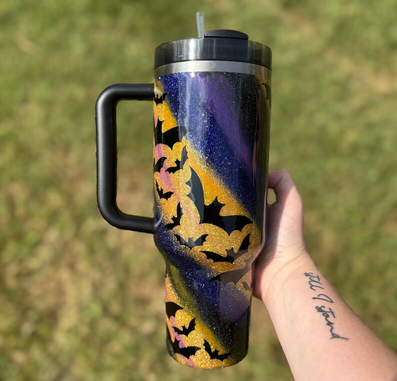 Puede incluir: Un vaso grande y aislado con asa y tapa negras. El vaso presenta un dise&ntilde;o brillante con un degradado de morado, dorado y azul. Siluetas de murci&eacute;lagos negros se superponen al brillo dorado, creando un tema de Halloween.