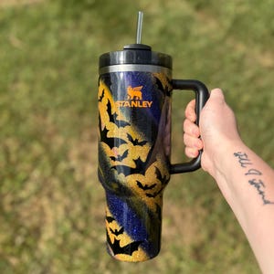 Puede incluir: Un vaso Stanley negro y dorado con asa, con un dise&ntilde;o de silueta de murci&eacute;lago. El vaso tiene tapa y pajita negras. El logotipo de Stanley es visible en naranja. El fondo es verde borroso.