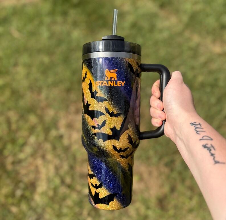 Puede incluir: Un vaso Stanley negro y dorado con asa, con un dise&ntilde;o de silueta de murci&eacute;lago. El vaso tiene tapa y pajita negras. El logotipo de Stanley es visible en naranja. El fondo es verde borroso.