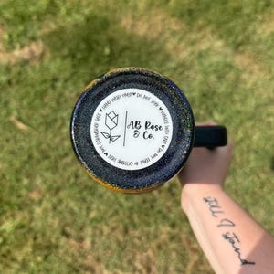 Puede incluir: Un vaso con asa negra y exterior brillante azul oscuro y dorado. La parte inferior del vaso presenta una etiqueta circular blanca con el texto "AB Rose & Co." e instrucciones de cuidado. El vaso est&aacute; sostenido por una persona.