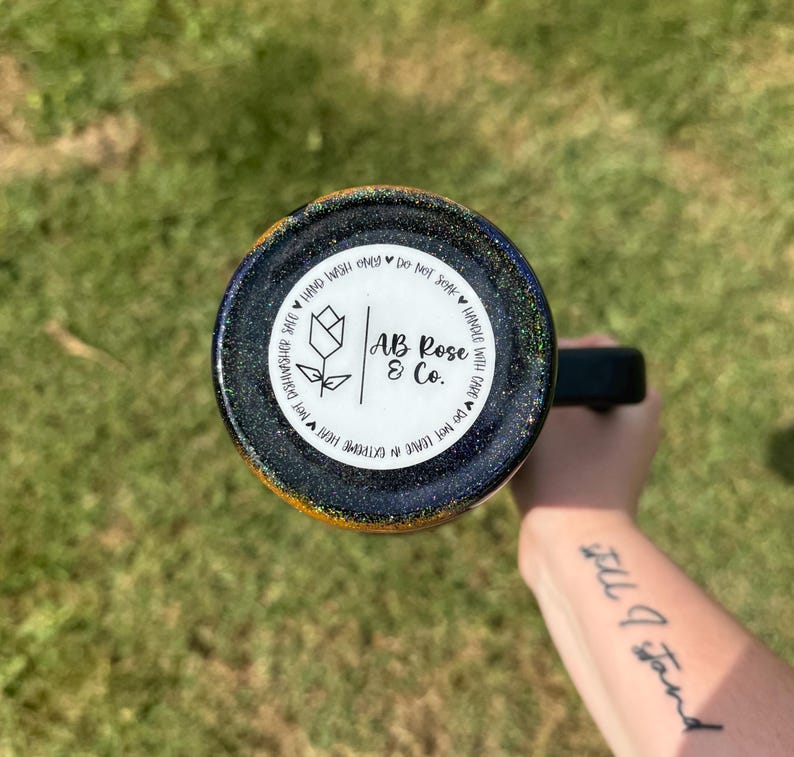 Puede incluir: Un vaso con asa negra y exterior brillante azul oscuro y dorado. La parte inferior del vaso presenta una etiqueta circular blanca con el texto "AB Rose & Co." e instrucciones de cuidado. El vaso est&aacute; sostenido por una persona.