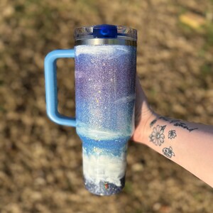 Puede incluir: Un termo cubierto de purpurina con asa y tapa azules. El termo presenta un dise&ntilde;o degradado con tonos azules, blancos y morados, que recuerda a un paisaje marino. El asa y la tapa son de color azul liso.