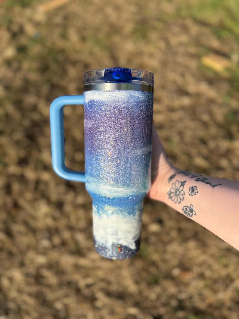 Puede incluir: Un termo cubierto de purpurina con asa y tapa azules. El termo presenta un dise&ntilde;o degradado con tonos azules, blancos y morados, que recuerda a un paisaje marino. El asa y la tapa son de color azul liso.