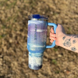 Puede incluir: Un vaso Stanley azul y blanco con purpurina, con asa y tapa azules. El vaso tiene un dise&ntilde;o degradado con purpurina blanca, azul y morada. La palabra "STANLEY" es visible en el lateral. El vaso est&aacute; dise&ntilde;ado para contener unos 887 ml.