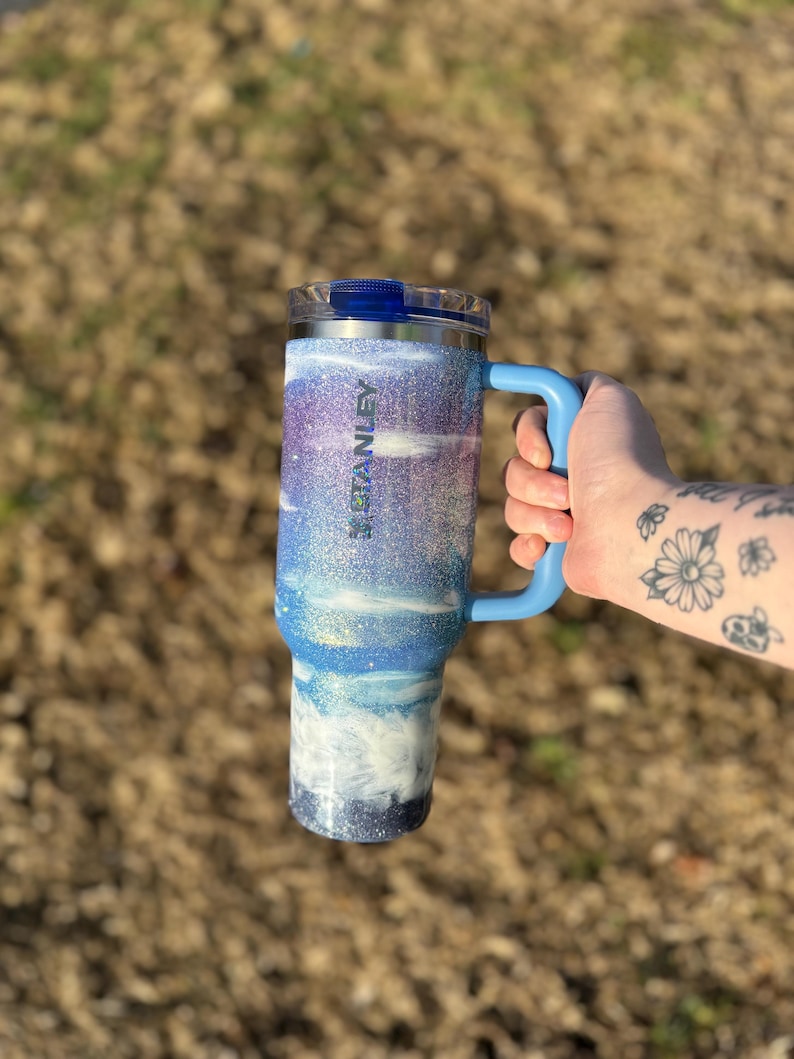 Puede incluir: Un vaso Stanley azul y blanco con purpurina, con asa y tapa azules. El vaso tiene un dise&ntilde;o degradado con purpurina blanca, azul y morada. La palabra "STANLEY" es visible en el lateral. El vaso est&aacute; dise&ntilde;ado para contener unos 887 ml.