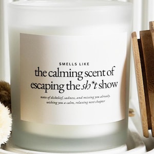 以下が含まれることがあります： 木製の蓋が付いた、すりガラスのキャンドル。キャンドルには「SMELLS LIKE the calming scent of escaping the sh*t show」と書かれた白いラベルが付いています。キャンドルは白いトレイの上にあります。