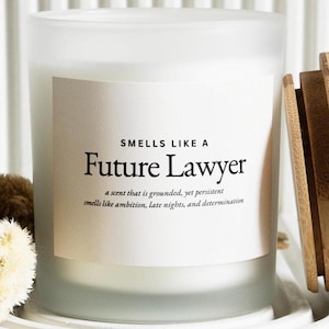 May include: A white frosted glass candle with a wooden lid. The label reads "SMELLS LIKE A Future Lawyer" with scent description. The candle is on a white, ridged surface. The candle is likely scented.