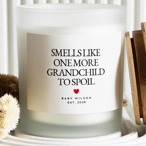 Pu&ograve; includere: Una candela bianca in un barattolo di vetro smerigliato con coperchio in legno. L'etichetta recita "Smells Like One More Grandchild To Spoil" con un cuore rosso e "Baby Wilson Est. 2026".