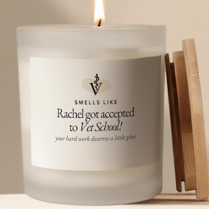 Könnte beinhalten: Eine brennende Kerze aus mattiertem Glas mit einem Holzdeckel. Die Kerze hat ein weißes Etikett mit dem Text "SMELLS LIKE Rachel got accepted to Vet School! your hard work deserves a little glow."