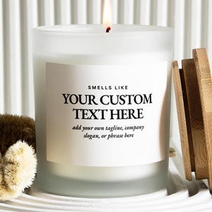 May include: A frosted glass candle with a lit wick and a bamboo lid. The white label features the black text: SMELLS LIKE YOUR CUSTOM TEXT HERE, add your own tagline, company slogan, or phrase here. The candle emits a warm, steady flame.